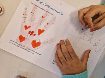 Kind beschriftet eine Hand auf einem Blatt Papier mit der Überschrift "Helfende Hände"