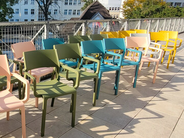 Stühle in grün, blau, gelb und rosa stehen aufgereiht auf einer Terrasse. 