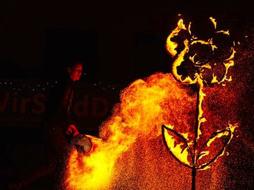 Eine Frau steht im Dunkeln, ein Lagerfeuer brennt. Eine große Blumenskulptur brennt.  