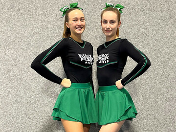 Zwei Cheerleader posieren mit ihren neunen Uniformen. Sie tragen jeweils einen grünen Minirock, ein schwarzes Oberteil und eine grüne Schleife im Haar. 