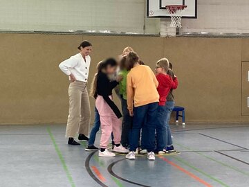 Kinder stehen zusammen in einer Gruppe. Eine Lehrerin steht daneben und beobachtet die Gruppe.