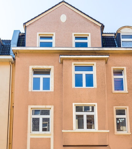 Fassade eines mehrstöckigen Wohnhauses mit orangefarbenem Putz und weißen Fensterrahmen. Das Gebäude hat mehrere Fensterreihen und ein Giebeldach.